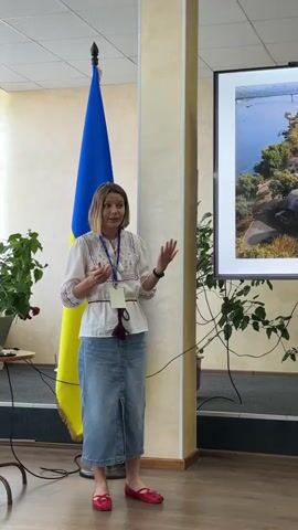 A woman stands in front of a Ukrainian flag, presenting a slideshow. She is wearing a white embroidered blouse, a denim skirt, and red flats. She has short blonde hair and is gesturing with her hands as she speaks. On the screen behind her is a picture of a bridge over a body of water, with trees and rocks in the foreground. The room appears to be a conference room or classroom, with a projector screen, plants, and a podium. The flag is prominently displayed, suggesting a formal event or presentation related to Ukraine. The lighting is bright and even, indicating an indoor setting during the daytime. The woman's expression is animated, suggesting she is engaged in her presentation. The overall scene depicts a professional or educational event.