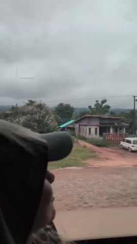 The video shows a person wearing a cap and hooded jacket, looking upwards and to the right. The person is positioned in the foreground, with their face partially visible. In the background, a rural landscape unfolds, featuring several houses with red roofs and visible vegetation, including trees. The ground appears to be dirt or gravel. The sky is overcast with clouds. The video appears to be shot from inside a moving vehicle, as evidenced by the blur and the angle of view.