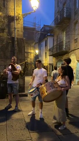 At twilight, a street performance unfolds in a cobblestone alleyway flanked by stone buildings with arched entrances and balconies. A man on the left plays bagpipes, wearing a white t-shirt and shorts. In the center, a man in a white t-shirt and shorts beats a small drum. To his right, a woman in a white crop top and a long skirt holds a large drum. Several people are dancing in a circle in the background, dressed in white t-shirts and dark shorts. A few spectators stand watching. The scene is lit by streetlights and ambient light from the buildings.