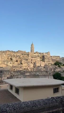 The video shows an upward-facing shot of a historic city built on a hillside, with a clear blue sky overhead. The buildings are constructed from light-colored stone and appear to be ancient, with many houses and structures clustered together on the sloping terrain. A prominent bell tower rises from the center of the city. In the foreground, a modern, flat-roofed building with windows is visible, partially obscuring the view of the older structures. A stone wall runs along the bottom of the frame. The lighting suggests it is either early morning or late afternoon, with the sun casting a warm glow on the stone. There are no people visible in the shot.