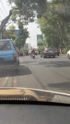 The video shows a street scene from the perspective of a vehicle. There are several cars and motorcycles on the road. The traffic appears to be stopped or moving slowly. There are trees and buildings on the sides of the road, and a billboard is visible in the background. The weather appears to be sunny.