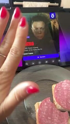 The video begins with a close-up of a person's hands with red nail polish, wearing a ring on the left ring finger. The hands are in the foreground, obscuring part of a tablet screen. The tablet displays a show titled "СЛЕД ЧИКАТИЛО" (The Trail of Chikatilo), identified as a new episode of season 4, a Russian thriller with a "18+" rating. The show's poster features a close-up of a person's face. Below the show title, there are icons for "Watch," "Add to Favorites," and other settings. In the lower right of the tablet screen, a purple gradient light is visible, possibly from another device or ambient lighting. In the bottom foreground, a glass plate with two open-faced sandwiches topped with sliced salami is visible. The background appears to be an indoor setting, possibly a table or desk, with some indistinct objects in the upper background. The hands move slightly during the clip, indicating interaction with the tablet or a gesture. The focus shifts slightly, and at 00:04, the person holds up two fingers.