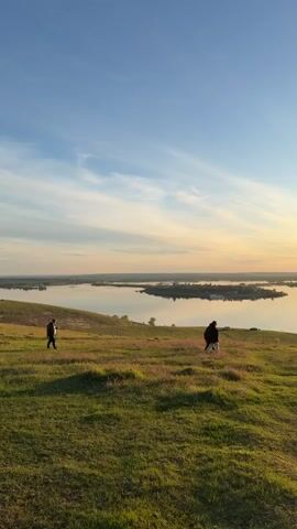 The video shows two adults and a child walking on a grassy hill with a large body of water in the background. The sky is blue with streaks of white clouds and a hint of orange from the setting sun. The water is calm and reflects the sky. The hill is covered in green grass with some patches of pink wildflowers. The people are walking away from the camera, and one of the adults is carrying a backpack.