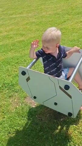 A young child with blond hair is sitting in a gray and white rocking toy outdoors on a grassy area. The child is wearing a dark t-shirt with a repeating white logo and light-colored shorts. The child's right hand is raised. The toy is tilted and appears to be in motion. The background is a sunny green lawn.