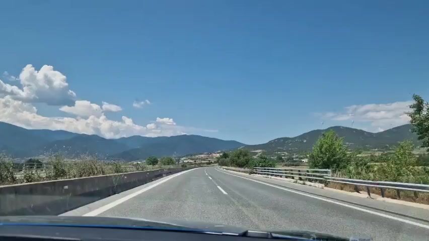 The video shows a view from inside a car driving on a highway on a sunny day. The road is straight and well-maintained, with white lines marking the lanes. On either side of the road, there are guardrails and some vegetation. In the distance, mountains can be seen under a clear blue sky with some scattered white clouds. The car appears to be moving steadily along the highway.