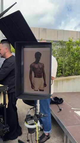 Here is a detailed description of the video:

The video shows a behind-the-scenes look at a photoshoot. The central focus is a large monitor, framed by a black hood to reduce glare, displaying a full-body image of a shirtless Black man with a muscular physique, wearing light-colored shorts. He is standing against a neutral-toned background.

Several people are present around the monitor. To the left, a man in a black shirt and black pants is partially visible, likely a crew member. A man in a white textured shirt and blue jeans stands to the right of the monitor, looking at his phone. Another man with a mustache and glasses peers over his shoulder at the monitor and its display of the model.

The monitor is mounted on a stand with visible cables and equipment attached, including a camera. The setting appears to be outdoors, on a raised platform or stage, with a building and green plants visible in the background.

The audio includes the indistinct voices of the crew, suggesting they are discussing aspects of the shoot, potentially related to the image displayed on the monitor.