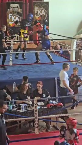 The video shows a boxing ring where two fighters are engaged in a match. One fighter is wearing black and yellow shorts with a blue headguard, while the other is in blue shorts and a red headguard. They are both equipped with gloves and shin guards. The ring is surrounded by ropes and has a blue mat. Around the ring, there are spectators watching the match. A table is set up near the ring with individuals appearing to be officials or judges, taking notes and monitoring the fight. There are water bottles and other equipment on the table. Banners with logos are hung in the background, indicating the event is organized or sponsored by specific organizations. The environment suggests a sporting event, specifically a boxing or martial arts competition.