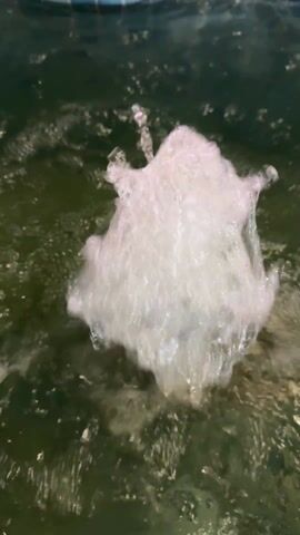 The video shows a water fountain in a pool. The water shoots up from a dark opening in the pool floor, creating a frothy, white spray against the green pool water.