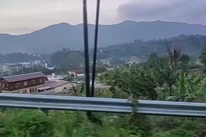 The camera is moving along a road, behind a metal guardrail, showing a view of a town or settlement nestled in a valley. The settlement includes buildings with dark roofs, interspersed with lush green vegetation. In the background, misty mountains rise against an overcast sky. Some utility poles are visible along the roadside.