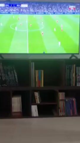 The video shows a television displaying a soccer match between Real Madrid (RMA) and Benfica (BEN). The score is tied 1-1, and the game is in the 47th minute. Players in white (presumably Real Madrid) and red (Benfica) are visible on the green field. The television is positioned on top of a brown bookshelf, which is filled with numerous books. The camera angle shifts slightly during the video.