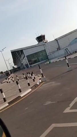 The video shows the exterior of an airport. The airport building is modern, with a large glass facade and a control tower. In front of the building is a large parking lot marked with yellow lines. There are numerous bollards painted with black and white stripes along the curb. The sign "Lubrik Construction Company" is visible. The sky is overcast, and the overall scene appears to be outdoors during the daytime. The video seems to be taken from a moving vehicle.