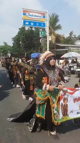 The video shows a group of people, including women and children, walking in a procession down a paved road. They are all wearing traditional, colorful costumes with ornate embroidery and headdresses that resemble feathers. The costumes are predominantly black with accents of green, red, and yellow. Some individuals are wearing headscarves. In the background, there is an Indomaret convenience store sign, trees, buildings, and parked motorcycles. The sky is visible and appears to be daytime. The procession is moving from right to left in the frame. Some people are carrying small flags.
