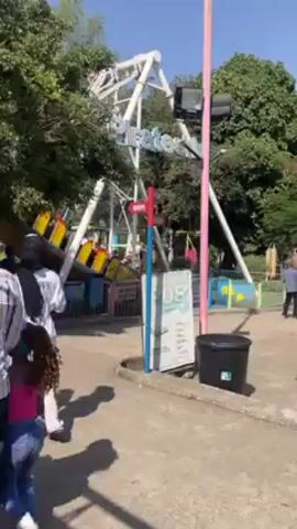 The video shows a fairground ride at an amusement park. The ride, named "Sea Dragon," features a swinging boat with passengers. There is a directional sign pointing towards "Shops" and "Arcade". People can be seen walking around the area, including a person wearing a green cap and a person in a blue shirt. The surroundings include trees, fences, and a trash can. The ride is in operation with people enjoying the experience.