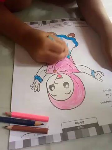 A close-up shot shows a child's hands coloring a picture of a cartoon girl wearing a pink hijab and blue dress. The girl is drawn in a slightly tilted position, with her head facing downwards. The child is using a light blue colored pencil to color the skirt of the dress. To the left of the coloring page, three colored pencils are visible: pink, brown, and dark blue. The coloring page has text at the bottom, with the words "Diriku" and "Topik" clearly visible. The background is out of focus but appears to be a floor with a light-colored tiled pattern.