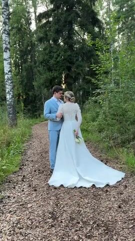 A couple, dressed in wedding attire, stands on a wooded path. The groom, in a light blue suit, faces the bride, who is wearing a white wedding dress with a long train and holds a small bouquet. They are looking at each other. The path is covered with wood chips and fallen leaves. Tall trees and foliage surround them, with a birch tree visible on the left. The lighting suggests it is daytime, possibly late afternoon.