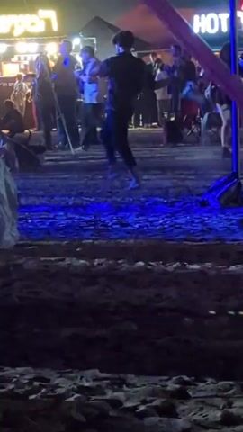 The video depicts a crowded outdoor scene at night, likely a festival or fair. People are milling around, some are dancing in the foreground on a sandy ground illuminated by blue lights. In the background, food vendors are visible under tents or stalls with signs like "HOT DOG" illuminated. The overall atmosphere is lively and bustling. A purple fabric hangs in the right side of the frame, adding to the ambiance.