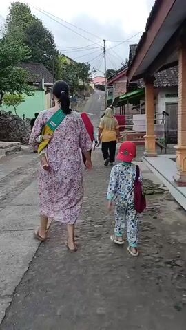 A rear view of four people walking up a slightly sloped, narrow paved road in a rural village. The person in the foreground on the left is an adult female wearing a long, floral-patterned dress and is barefoot. She has a baby in a sling across her chest. To her right and slightly ahead is a child, likely a boy, wearing a blue and white patterned tracksuit and a red baseball cap. He is carrying a maroon bag over his right shoulder. Further up the road are two other individuals, both women, walking ahead of the child. The person closer to the child is wearing a red hijab and a blue top. The person furthest ahead is wearing a yellow top and a light-colored hijab. The road is lined with houses and some vegetation. The sky is overcast. A motorcycle is visible in the distance further up the road.