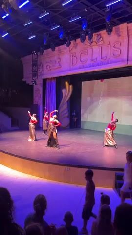 A video shows a stage performance at the Bellis Deluxe Hotel. Several female dancers in gold and red costumes perform on a lit stage. The stage has a large screen in the background displaying a chain-like pattern. The audience is visible in the foreground, with several children and adults watching the performance. The lighting is dim and theatrical.