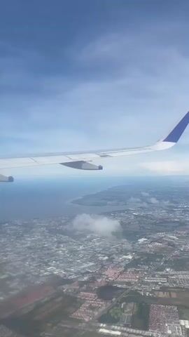 The video shows a view from an airplane window, looking out over a city near a body of water. The wing of the plane is visible in the foreground, white with a blue tip. The sky is mostly clear blue with some wispy clouds. Below, the city stretches out with buildings and roads visible. Beyond the city, there's a large body of water with a visible shoreline. Some scattered clouds are also visible over the city. The overall scene is bright and sunny.