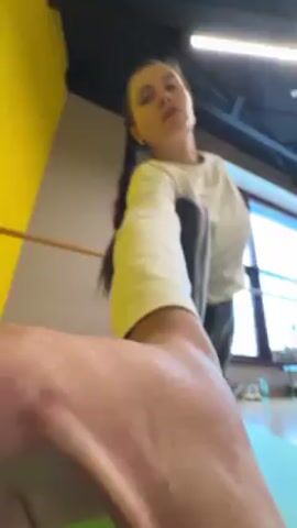 A young woman is filmed from a low angle in what appears to be a dance studio or gym. She's wearing a white long-sleeved shirt, dark leggings, and white socks, with her hair in a braid. She stands on a green yoga mat, in a pose stretching her leg, her arms held out. Behind her, there is a barre along the wall, a window, and another yoga mat on the floor. The room has a yellow wall in the background and a dark ceiling with visible fixtures.