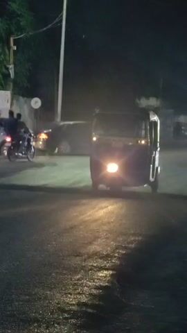 The video shows an auto-rickshaw driving on a wet, dark road at night. The auto-rickshaw's headlight is on, illuminating the road ahead. In the background, a person is riding a motorcycle. The road appears to be a street with some buildings and trees lining the sides. The overall atmosphere is that of a city street at night, possibly during or after rain.