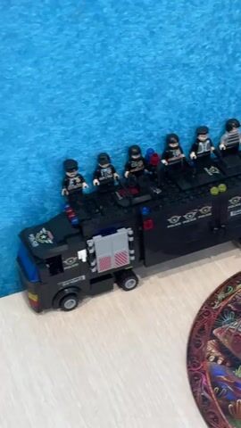 The camera pans to the right from a LEGO police truck with LEGO figures on top to a round puzzle of a turtle. The background is a blue, textured wall. The objects are sitting on a light-colored wooden table. There is a thin, silver pole directly behind the puzzle.