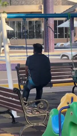 A man is seated on a swing set in an outdoor area, likely a resort or hotel playground, during the daytime. He is seen from behind, wearing a dark patterned shirt, blue jeans, and sneakers. A light blue band, possibly underwear, is visible above his waistband. He is looking towards a swimming pool area with lounge chairs and umbrellas, and further back, a building with balconies and glass windows. In the foreground, another wooden bench is visible, along with green and yellow children's play equipment. The swing set has a white A-frame structure and blue chains. The overall setting suggests a leisure environment in Al Fiqait, United Arab Emirates. The man is still and appears to be observing the surroundings rather than actively swinging.
