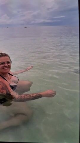 The video shows an eye-level view of a calm body of water. A person is seen from the knees up, their arms submerged in the water. They have visible tattoos on their right arm. Several small fish swim near the surface of the water. In the distance, land can be seen against a cloudy sky. The water is shallow and clear enough to see the sandy bottom. The overall impression is of a calm, natural setting.