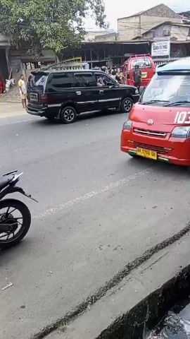 A black SUV and a red van are stopped on a paved road. Several people stand on the sidewalk to the left of the road, some of them looking towards the vehicles. A motorcycle is parked on the left side of the road, facing away from the camera. A yellow truck with the words "NADIA GROP" on its side is moving on the road behind the black SUV. The scene appears to be in a commercial area with buildings visible in the background.