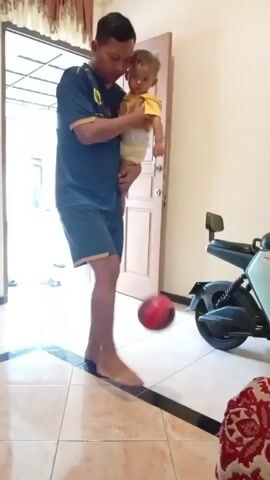 A man in a dark blue t-shirt and dark blue shorts holds a young child in his arms while inside a house. The man is balancing a red ball on his knee, but he loses control of it. The room has tile floors, a light blue motor scooter, and a door leading to the outside.