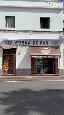 The video shows a storefront for a bakery named "HORNO DE PAN" which translates to "Bread Oven". The facade of the building is white with gray trim at the bottom. Two windows are visible on the upper floor, one with closed white curtains and the other with open green shutters. The storefront is divided into two sections. The left section has a wooden door with a metal security gate and a display window showing various loaves of bread. The right section has a glass door with an awning above it. This awning, as well as the awning over the left entrance, are a dark brown color and have the bakery's name and logo on them. The logo depicts a baker at an oven and the words "HECHO AQUI" which means "Made Here". Inside the right section, there are shelves with bread and pastries. A yellow "Wet Floor" sign is visible inside the right entrance. The ground in front of the bakery is paved with dark asphalt and a pinkish-red border along the sidewalk. A shadow from the building falls across the facade. A pink sign that reads "BISERVICUS" is mounted on the wall to the right of the left entrance.
