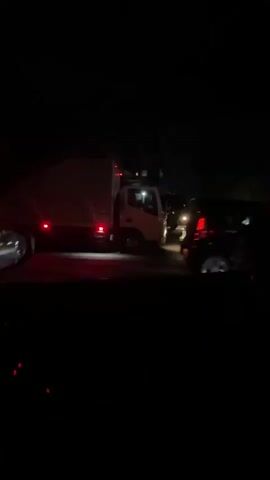 The video captures a night scene on a road. A white box truck with red taillights and additional red marker lights on its side is the primary subject. The truck is initially seen in the foreground, facing away from the camera and slightly to the right, then proceeds to drive away. Its brake lights illuminate intermittently. Other vehicles' headlights and taillights are faintly visible in the dark background, suggesting traffic or parked cars further down the road. A white and orange traffic cone is briefly visible in the lower left foreground, seen through what appears to be a car window. The setting is very dark, with artificial lights from the vehicles and distant streetlights providing the only illumination. The video was captured at night in what is identified as Sri Jayewardenepura Kotte, Sri Lanka. No discernible text is visible.