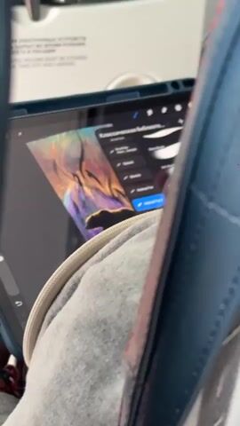 A person sits on an airplane, holding a tablet device and a stylus. The tablet screen displays a digital painting of a bear in a forest setting, with various editing tools visible. The person is using the stylus to interact with the tablet, likely editing the artwork. The focus is on the tablet and the person's hands, with the airplane cabin visible in the blurred background.