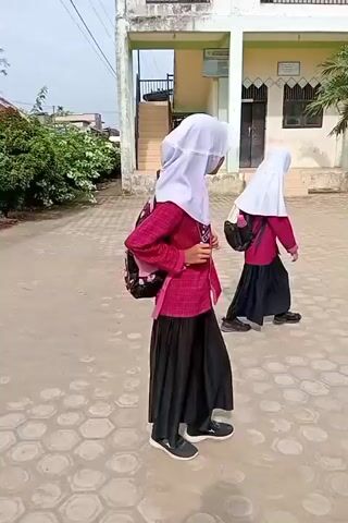 Two young girls in hijabs and long black skirts are walking away from the camera. The girl in the foreground has a pink and black backpack with a floral pattern. She is wearing a pink shirt. The girl behind her has a black backpack and is wearing a similar pink shirt and black skirt. They are walking on a paved area outside a building that appears to be a school. The building is light-colored with some green accents. There are palm trees and bushes in the background. The pavement has a honeycomb pattern. The lighting suggests it is daytime, possibly mid-morning or afternoon, with bright sunlight casting shadows. The video is shot from a slightly low angle, following the girls.
