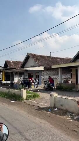 The video shows a street scene with a single-story house. In front of the house, there is a clothesline with clothes hanging on it. A person is riding a motorcycle on the right side of the frame, moving away from the camera. The house has a tiled roof and is made of brick and painted walls. There are power lines running overhead. The ground in front of the house is paved, and there is some vegetation on the left side of the frame. The sky is clear and blue with some clouds. The overall impression is a residential area in daylight.