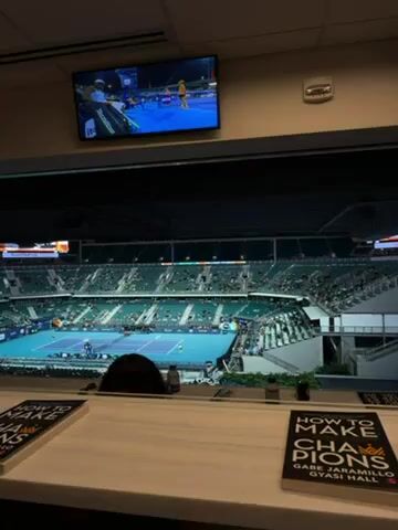 The video shows a tennis match in a large stadium with many spectators. The view is from a high vantage point, possibly a suite.  A television screen above the main view shows the match from a closer angle. There are two books titled "How to Make Champions" on the table in front of the viewer. The tennis court is blue, and players are visible in white and yellow outfits. The stadium is well-lit, and the stands are filled with people watching the game. A person's dark hair can be seen in the lower center of the frame.