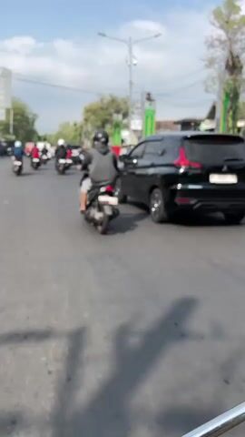 A black SUV is driving down a paved road. Several motorcycles are also on the road, some in front of and some behind the SUV. One motorcyclist in the foreground is wearing a dark hoodie and riding a lighter-colored motorcycle. The environment appears to be a city street with buildings and trees lining the sides of the road. The sky is partly cloudy.