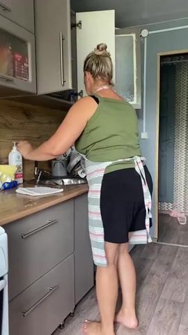 A woman in a green tank top, black shorts, and a striped apron stands in a kitchen, facing away from the camera. She has her hair pulled back in a bun and is barefoot. She is reaching into an open cabinet above a sink. The cabinet contains various bottles and containers. The kitchen has gray cabinets, a wooden countertop, and a tiled floor. A sink with dishes is visible, along with a bottle of dish soap and a yellow sponge. The background shows a doorway with a patterned curtain and a wall.