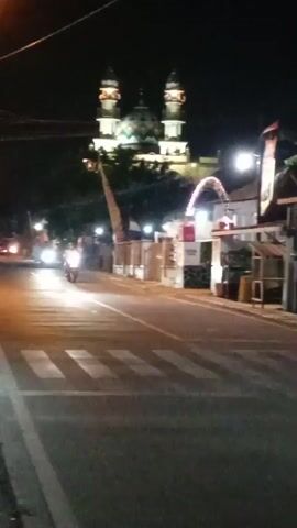The video shows a street at night with a mosque in the background. A motorcycle is riding down the street, and there are buildings on the right side of the road.