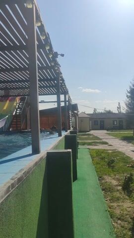 The video shows an outdoor scene on a sunny day. In the foreground, there is a swimming pool with clear blue water. On the right side of the pool, there is a green surface, possibly astroturf, and a paved pathway. Beyond the pool, there is a wooden structure with a slatted roof and lights. Further in the background, there are buildings, trees, and a clear blue sky with some clouds. The sun is visible in the upper left corner of the frame. The camera angle is tilted, making the scene appear to be on its side. There is a person partially visible in the swimming pool.