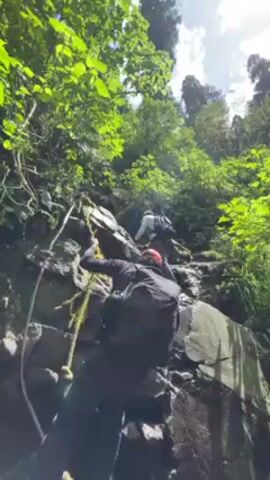 A low-angle shot shows a person in dark clothing and a black backpack ascending a rocky, green hill using a rope.  The rope appears to be a yellowish-brown color. Another person is visible higher up the hill.  The terrain is rocky and wet, possibly near a waterfall or stream.  The surroundings are lush with green vegetation and trees under a bright sky with scattered clouds. The sun is shining, highlighting the green foliage. The scene suggests a challenging outdoor adventure.