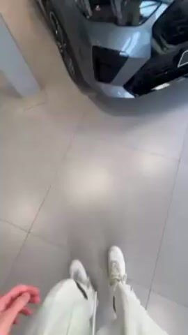 The video shows a showroom of BMW cars. There are several models of BMWs on display, including SUVs and sedans. The showroom is well-lit and has a modern design with white tile flooring. There is also a seating area with brown chairs and white couches. Outside the large windows, the street and traffic can be seen. The camera moves around the showroom, showing different angles of the cars.