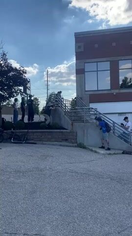 A group of young men are gathered outdoors near a brick building. Several of them are on a set of concrete stairs with metal railings. One person is on a scooter at the bottom of the stairs and appears to be preparing to do a trick. Another person on a scooter is in the foreground, looking towards the stairs. Several other individuals are standing or sitting around the area, some holding phones. The sky is blue with scattered clouds. There is a parking lot in the foreground and a street with cars in the background.