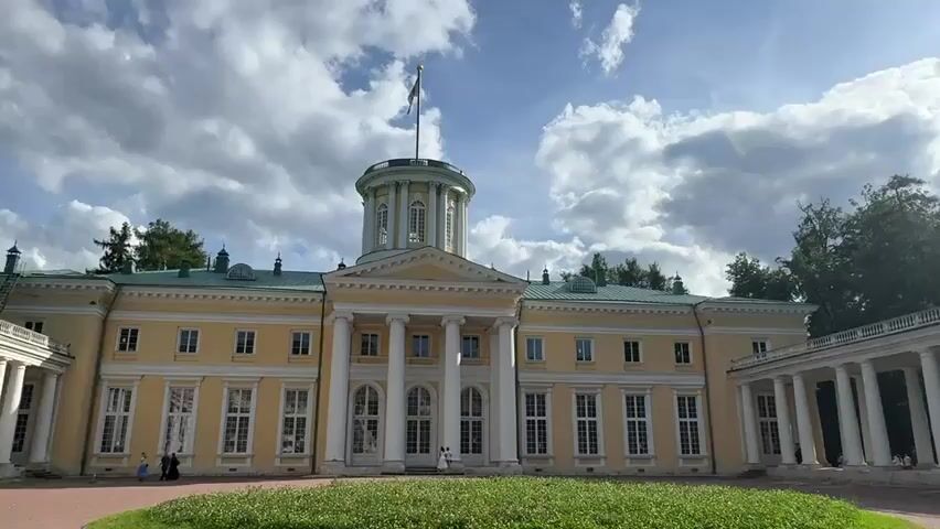 The video shows a wide, low-angle exterior shot of a large, pale yellow neoclassical building with a prominent central rotunda topped by a flagpole. The building features a grand portico with tall white columns, multiple windows, and green roofing. To the right of the main building, a long, colonnaded walkway extends outwards, also supported by white columns. The ground in front of the building is a reddish-brown paved area with a circular patch of green grass in the foreground. In the background, lush green trees are visible. The sky is partly cloudy with patches of bright sunlight. A few people are visible in the distance near the building and in the colonnaded walkway. Scaffolding is present on the left side of the building, suggesting ongoing construction or restoration work.
