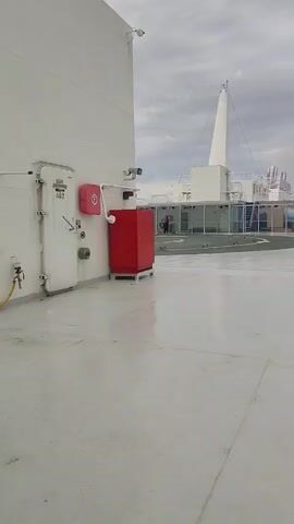 A person walks along a white deck of a ship. The person is wearing a dark jacket and pants. The person is walking away from the camera towards a white railing. Beyond the railing is a body of water and a cloudy sky. There is a red life preserver hanging on the railing. To the left of the railing are several trash cans. The deck is wet, reflecting the cloudy sky. In the background, there is a white superstructure with large windows. There is a tall white mast extending upwards from the superstructure. The deck is clean and appears to be well-maintained. There is a red fire extinguisher cabinet mounted on the white wall of the ship, with a fire hose coiled inside.