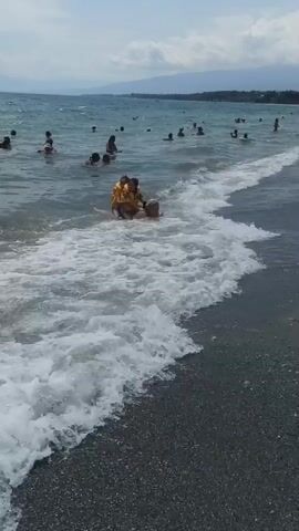 The video depicts a crowded beach with many people swimming and wading in the ocean. The water is choppy with waves breaking on the shore. In the foreground, two children in yellow life vests are sitting on a dark-colored object, possibly a float or small raft, in the shallow water. Waves are crashing around them. Further out in the water, numerous individuals of various ages are visible. The beach itself appears to be covered in dark pebbles. In the background, a mountain range is visible under a cloudy sky. A man wearing a cap and dark shorts walks away from the camera on the beach in the lower right portion of the frame.
