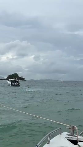 A boat is partially submerged in choppy green water. It is angled with the stern higher out of the water than the bow. Several people are visible on the deck of the submerged boat. Another boat is visible in the distance to the right, and a small, tree-covered island is to the left of the submerged boat. The sky is overcast and cloudy. A rope extends from the foreground boat towards the submerged boat.