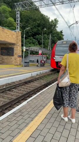 The video shows a train station platform with a red and gray passenger train approaching. A woman wearing a yellow t-shirt and patterned pants stands on the platform, holding a black plastic bag. Another person, partially visible, stands to her right. The platform is paved with gray bricks and has a yellow line marking the edge. In the background, there is a yellow brick building, train tracks, overhead power lines, and lush green trees on a hillside. The train has the Russian Railways logo visible.