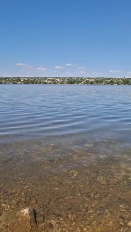The video shows a clear blue sky above a calm body of water. The water is shallow near the shore, revealing a rocky bottom. A stone is dropped into the water, creating ripples that spread outwards. In the distance, a town or village can be seen along the shoreline. The overall scene is peaceful and serene.