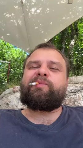A close-up, selfie-style video shows a man with a beard smoking a cigarette. He is outdoors, with lush green trees and a white umbrella visible in the background. The man takes a drag from the cigarette, exhales, and then opens his mouth wide, revealing the cigarette in his mouth. The video is short and focuses solely on the man and his action.